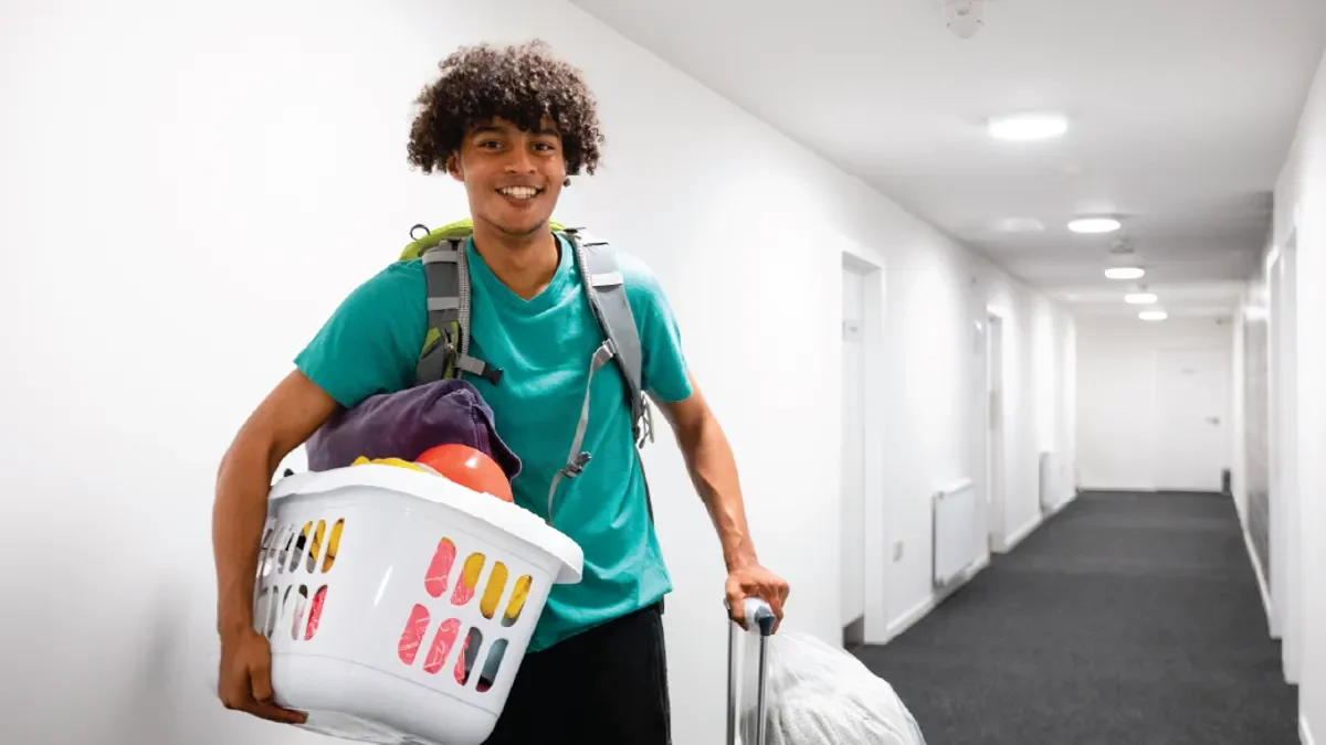 Smiling person with a backpack carries a laundry basket and pulls a large white bag through a hallway.